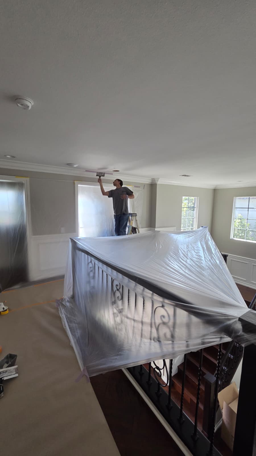 Painter on a ladder working on an interior ceiling above a covered stair railing